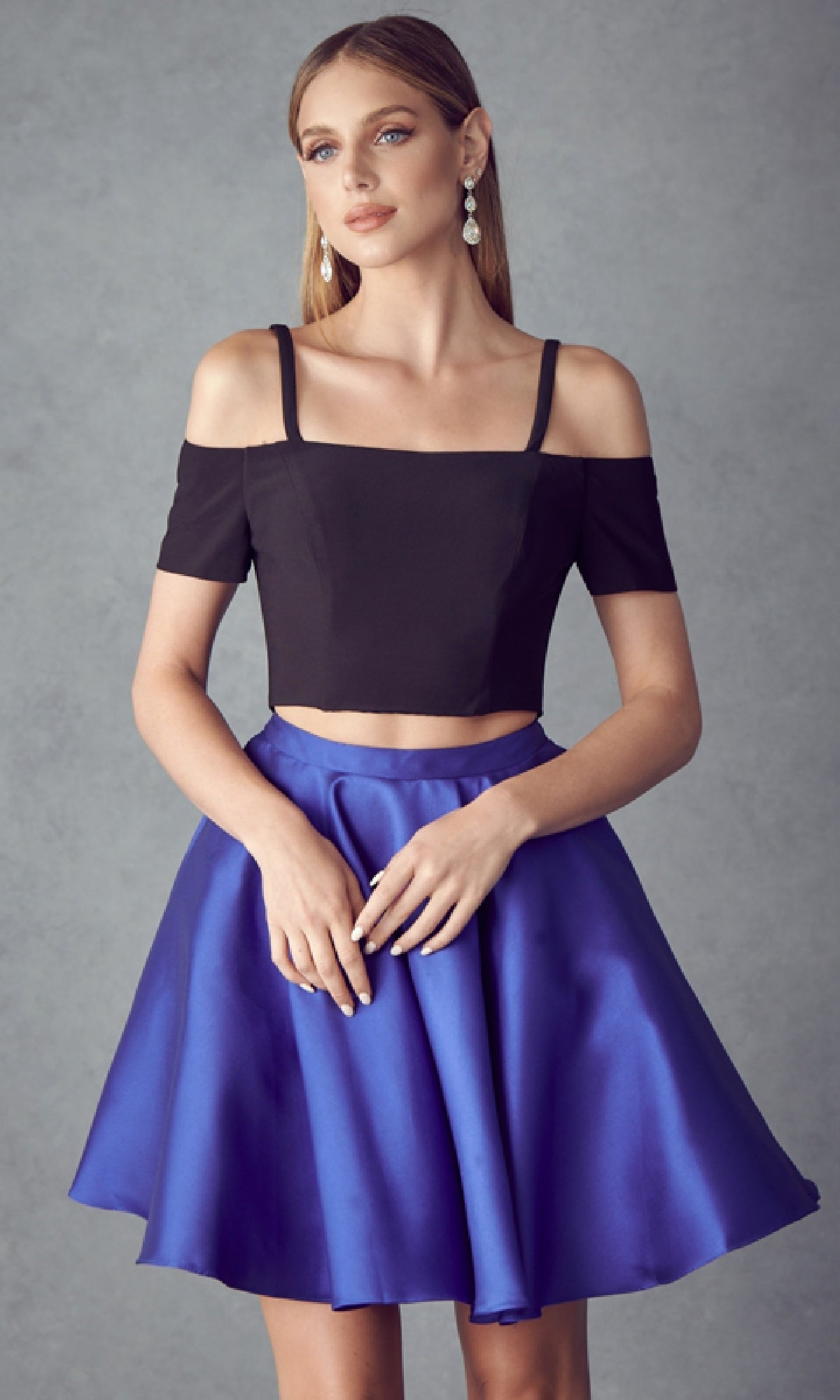 A woman stands against a gray background wearing the Short Party Dress: Juliet 809—a black off-the-shoulder crop top with a shiny blue A-line skirt—paired with drop earrings and straight hair.